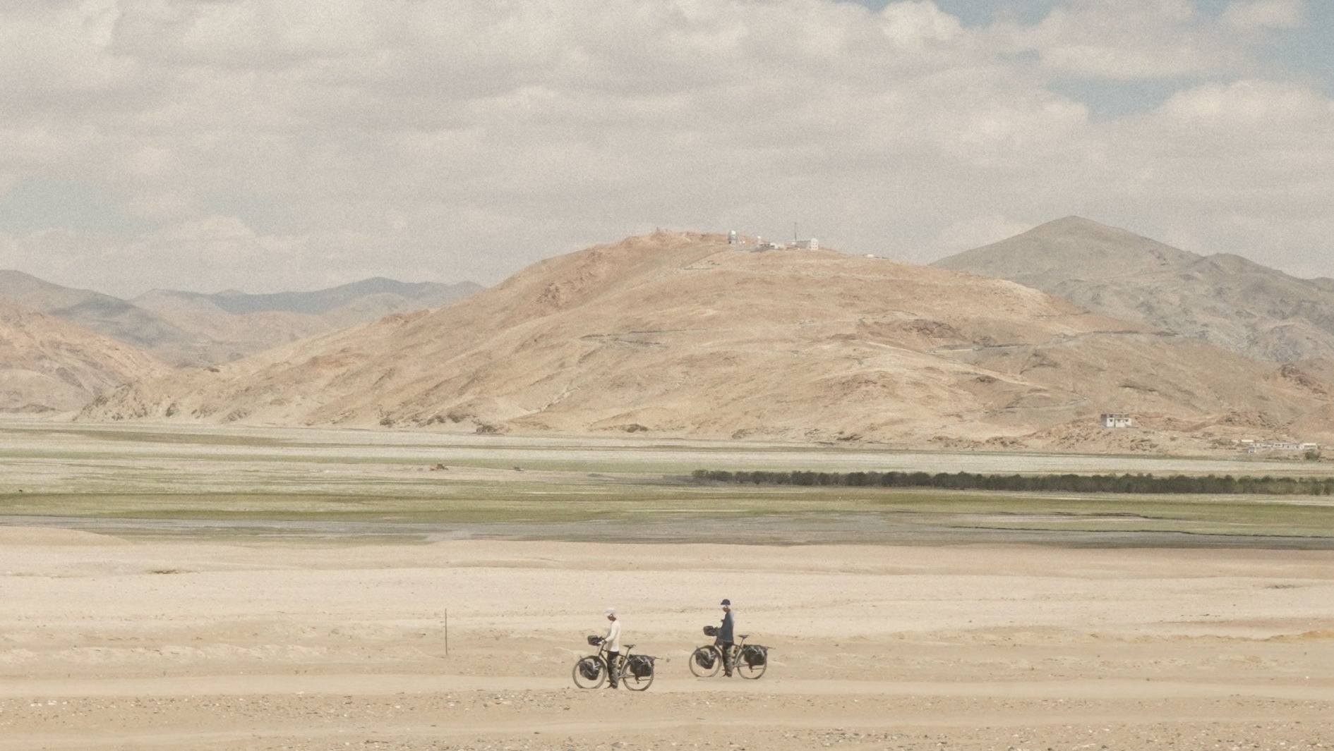 Deux cyclistes et leurs vélos chargés dans la plaine du Ladakh, face aux montagnes — visuel adapté de l'affiche du film